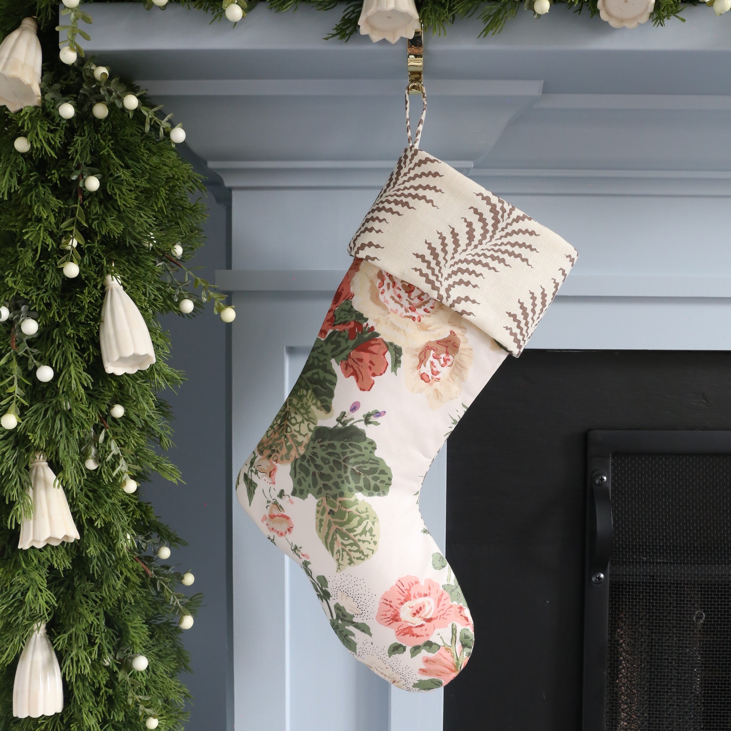 Floral-patterned Christmas stocking hanging on a fireplace with greenery and lights.