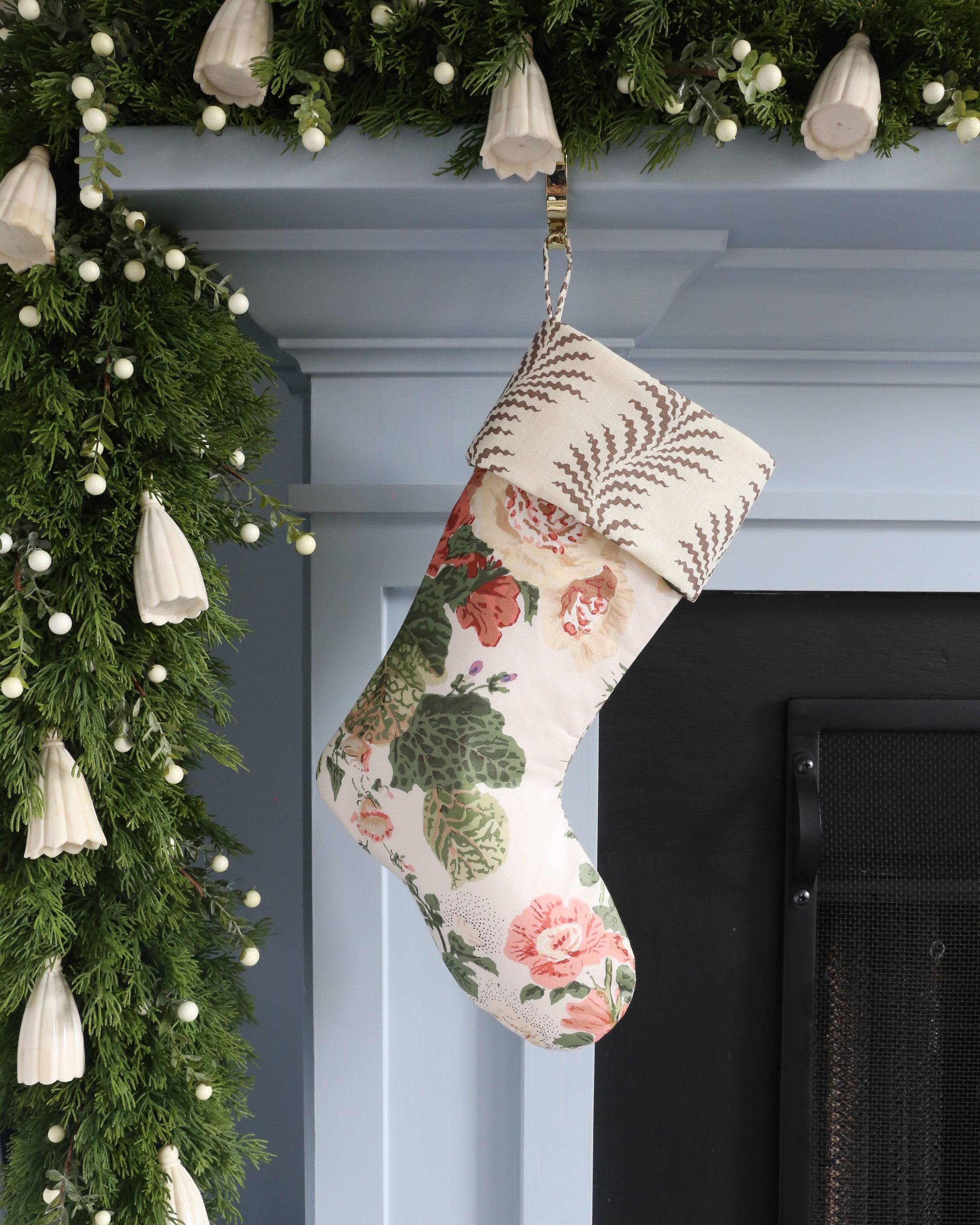 Floral-patterned Christmas stocking hanging on a fireplace with greenery and lights.